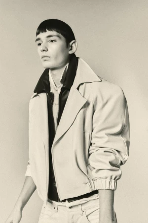 Portrait of a young adult with a bowl cut, wearing a light-colored jacket, collared shirt, and jeans against a neutral background.