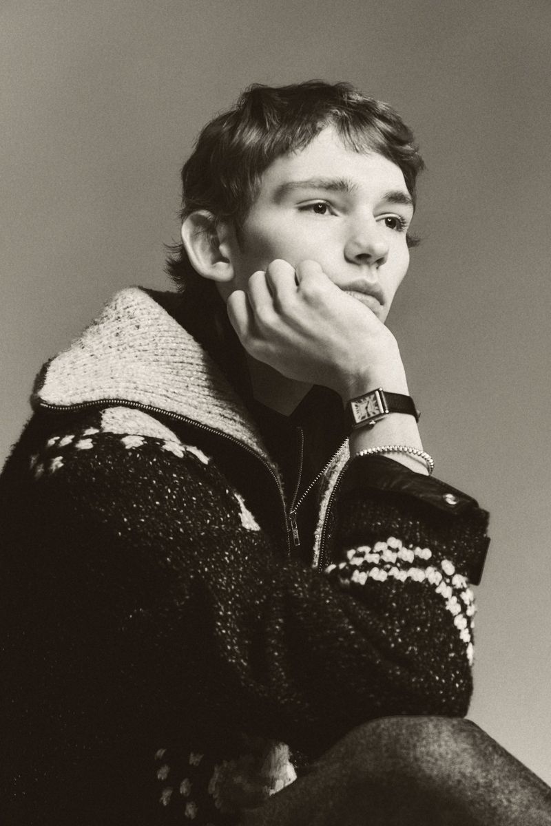 Portrait of a young adult with short brown hair, wearing a knitted zip-up jacket and a watch, resting their chin on their hand.