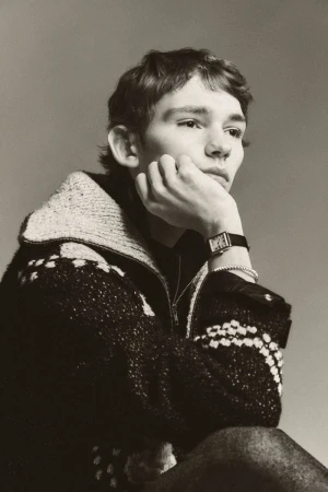 Portrait of a young adult with short brown hair, wearing a knitted zip-up jacket and a watch, resting their chin on their hand.