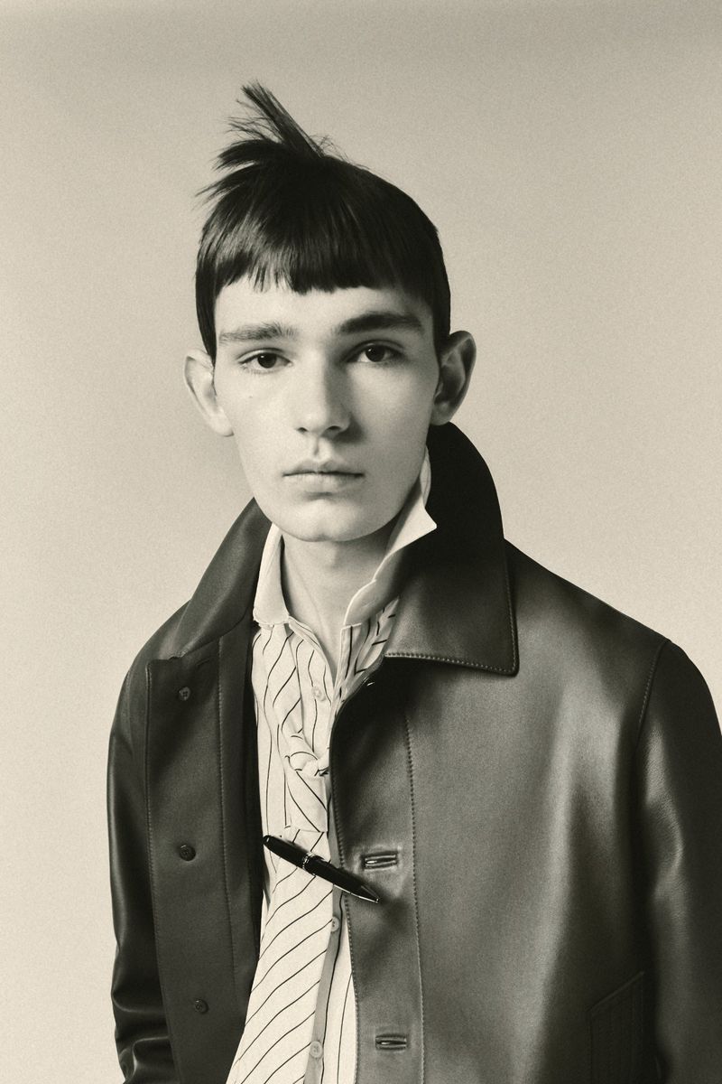 Monochrome portrait of a young adult man with an unusual hairstyle wearing a striped shirt and leather coat against a light background.