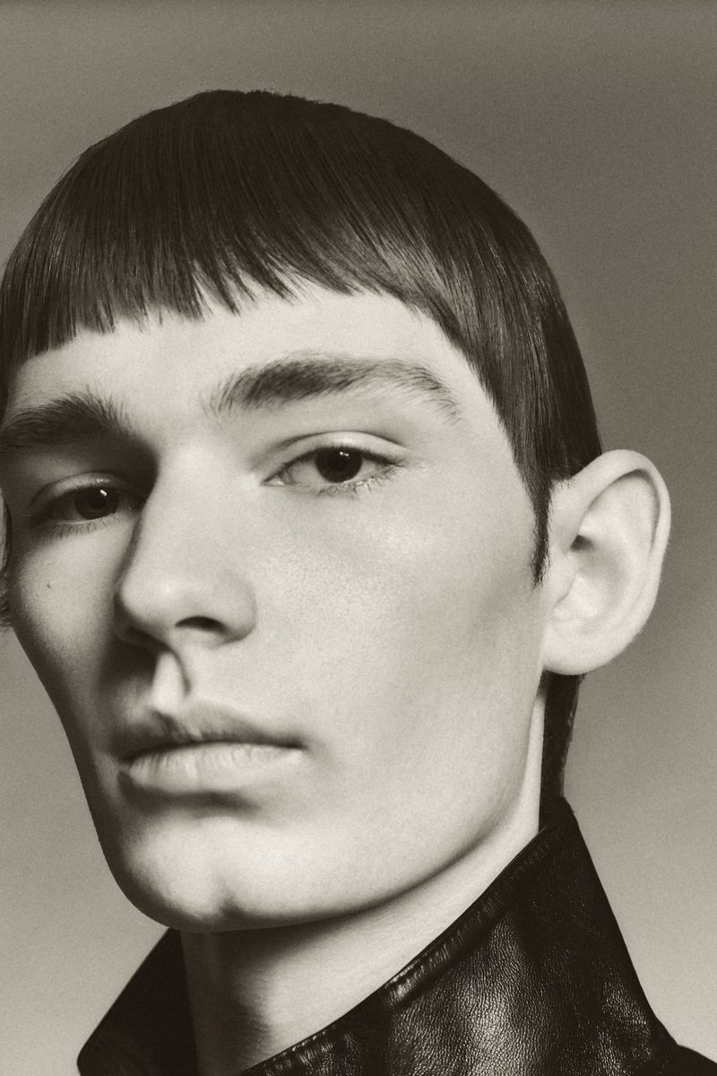Close-up portrait of a young man with straight bangs wearing a dark collared leather jacket against a neutral studio background
