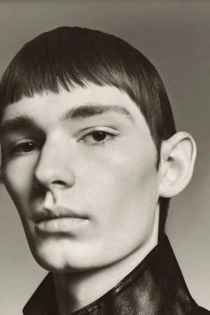 Close-up portrait of a young man with straight bangs wearing a dark collared leather jacket against a neutral studio background