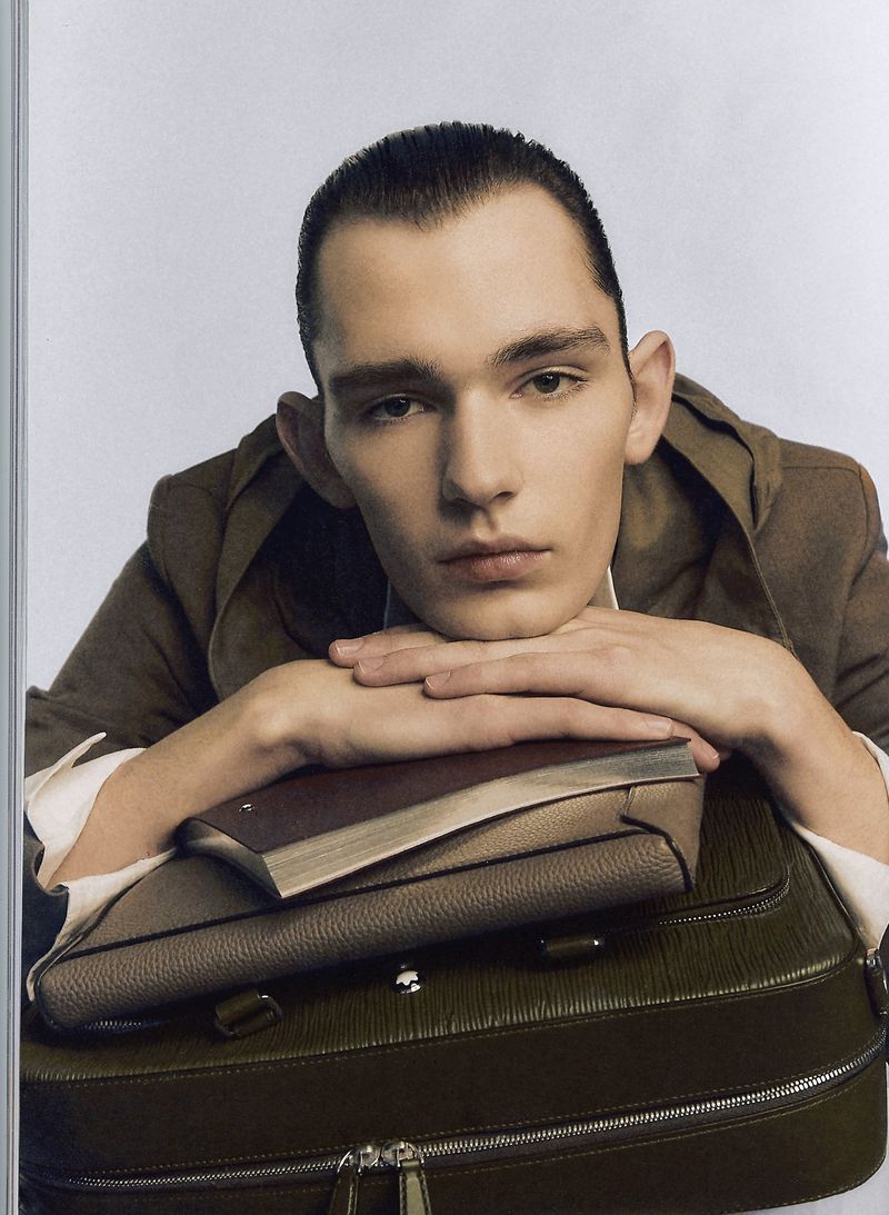 Portrait of a young adult man resting his chin on his hands atop a dark olive briefcase and a closed book against a white background.