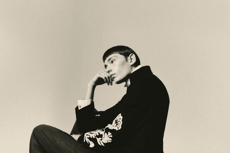 Model with short dark hair in profile, wearing an embroidered dark jacket, resting head on hand against a neutral studio background