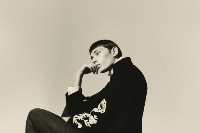 Model with short dark hair in profile, wearing an embroidered dark jacket, resting head on hand against a neutral studio background