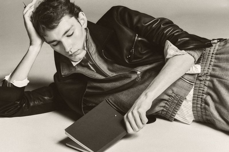 Young adult laying with a book, wearing a leather jacket and textured pants in a studio setting, looking away in sepia tone.