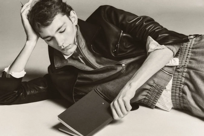 Young adult laying with a book, wearing a leather jacket and textured pants in a studio setting, looking away in sepia tone.