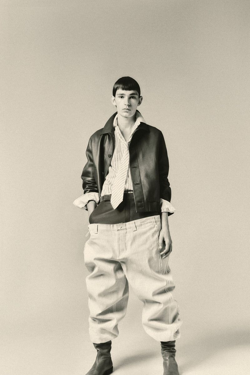 Young adult wearing a black leather jacket, striped shirt, khaki pants, and ankle boots in a full-length studio shot.