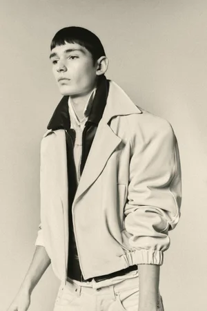 Young male model with dark bowl cut hair wearing an off-white bomber jacket with black collar against a neutral studio background