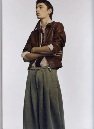Young man with arms crossed wearing a brown leather jacket, striped shirt, tie, and grey pleated trousers against a plain wall