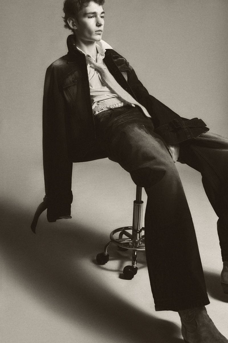 Young male model seated on a rolling stool wearing a dark jacket, collared shirt, and relaxed dark trousers against a neutral background.