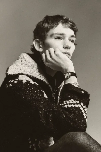 Young man with short textured hair resting his chin on his hand, wearing a patterned knit zip-up cardigan against a neutral grey studio backdrop.