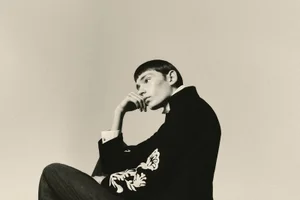 Model with short dark hair in profile, wearing an embroidered dark jacket, resting head on hand against a neutral studio background