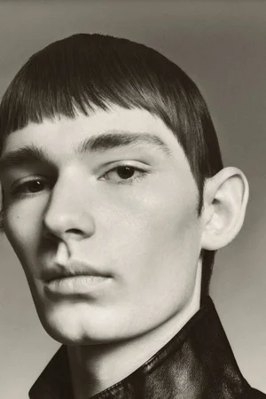 Close-up portrait of a young man with straight bangs wearing a dark collared leather jacket against a neutral studio background