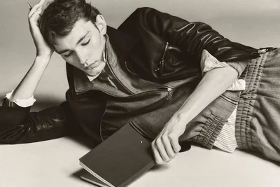 Young man in a black leather jacket and grey trousers lying on a light studio background holding a notebook