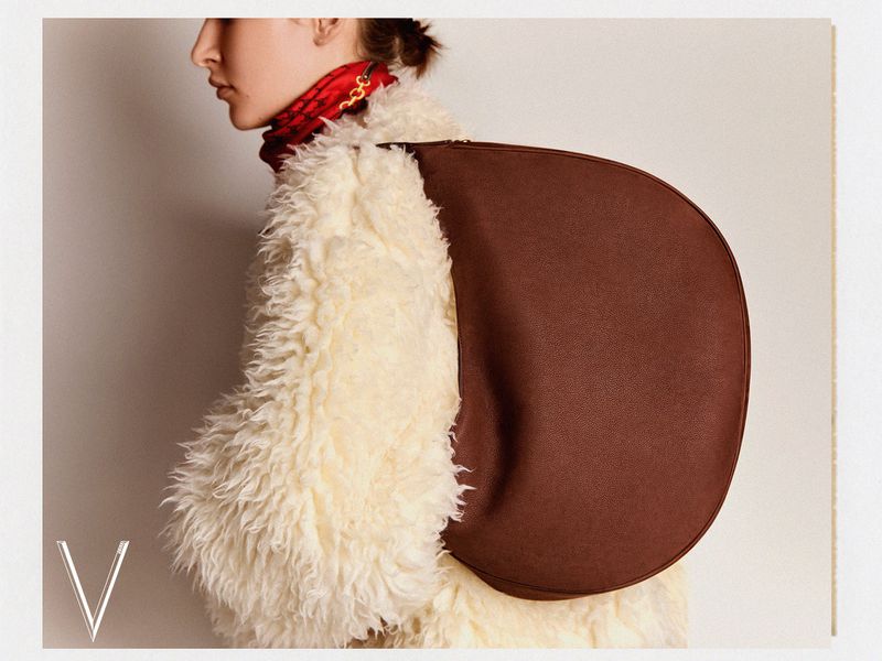 Fashion shot featuring a woman in a cream-colored, fuzzy coat with a brown leather backpack, side view on an off-white background.