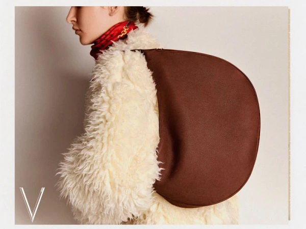 Model in fluffy white coat wearing a brown crescent shoulder bag against a plain off-white background