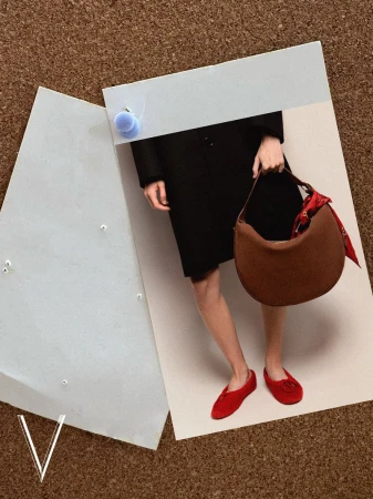 Close-up fashion editorial shot of a person wearing a black coat, red shoes, and holding a brown shoulder bag against a corkboard background.
