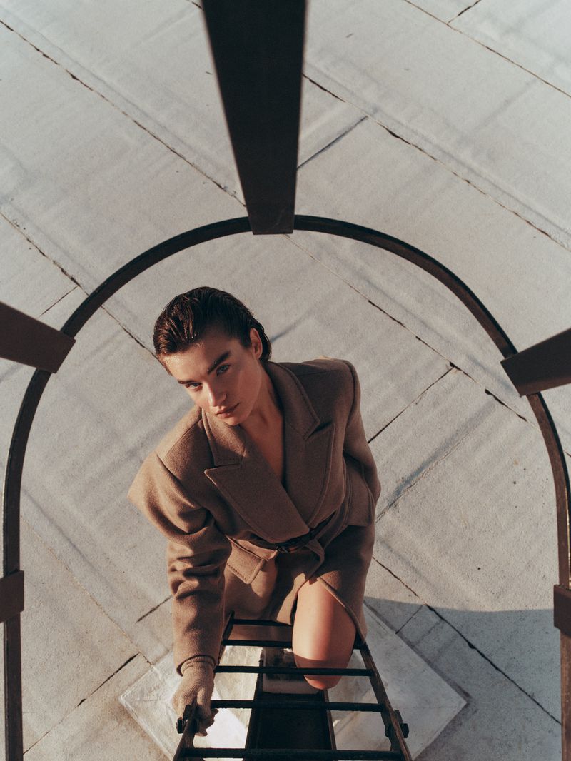 Eye-level shot of a woman in a brown belted jacket climbing a gray metal ladder against a white ceiling backdrop, fashion editorial.