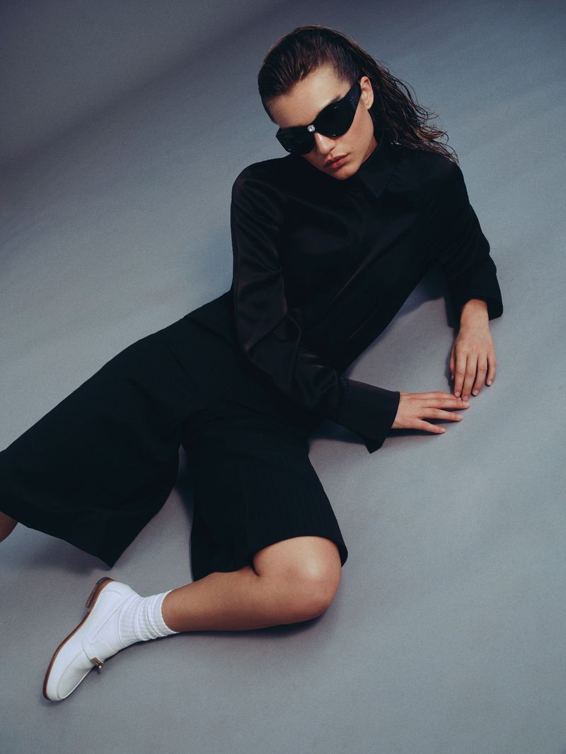 Model Meghan Roche wearing black clothing and sunglasses seated against a grey studio backdrop for Harper's Bazaar Greece