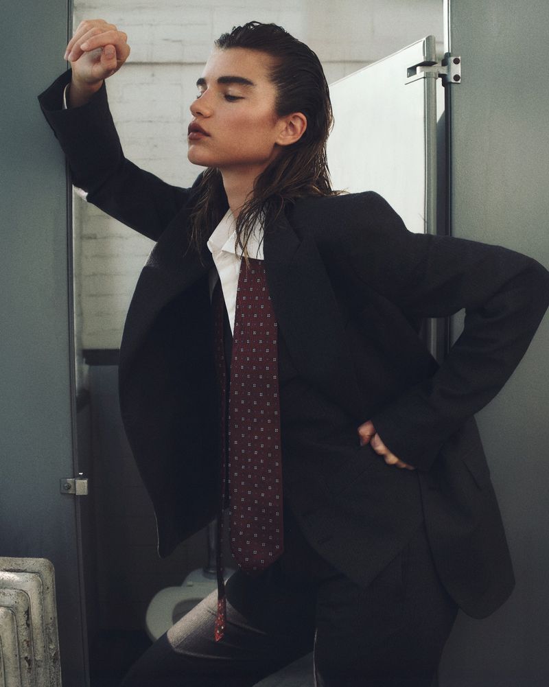 Woman in a suit leaning against a door in a bathroom, wearing a white button-down shirt with a dark tie and black suit jacket and pants.