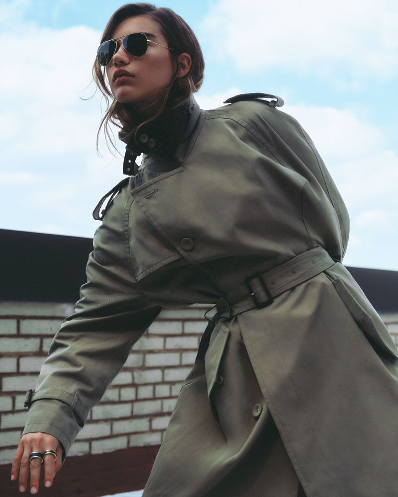 Adult female in a trench coat and sunglasses stands on a rooftop against a sky backdrop, creating a high-fashion editorial image.