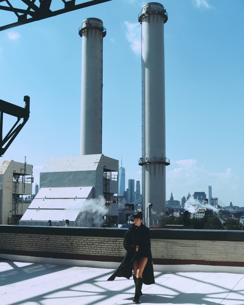 Fashionable woman in a black trench coat poses on a rooftop with industrial towers and a distant city skyline on a sunny day.