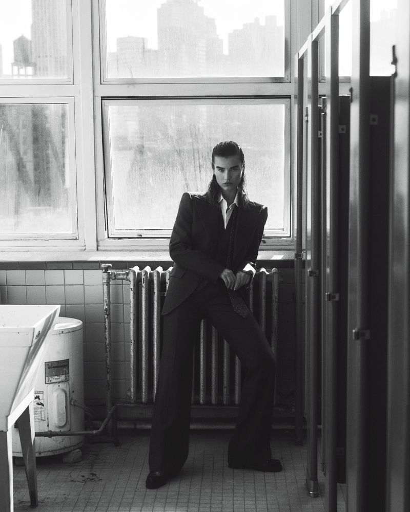 A model poses in a black suit inside a tiled public restroom in a black-and-white editorial fashion photograph with window light.