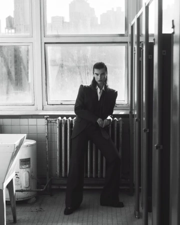 Model in dark pantsuit standing before large windows and a radiator in a black and white fashion editorial photograph.