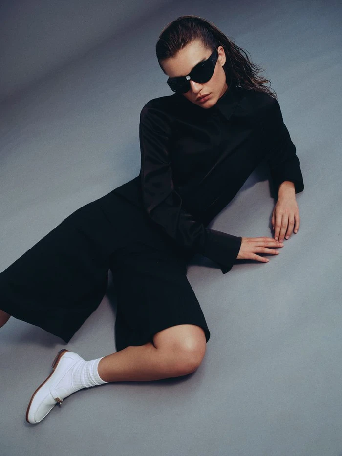 Model Meghan Roche wearing black clothing and sunglasses seated against a grey studio backdrop for Harper's Bazaar Greece