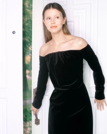 A fair-skinned woman in a black off-the-shoulder dress holds the handle of a white door leading to an outdoor landscape.
