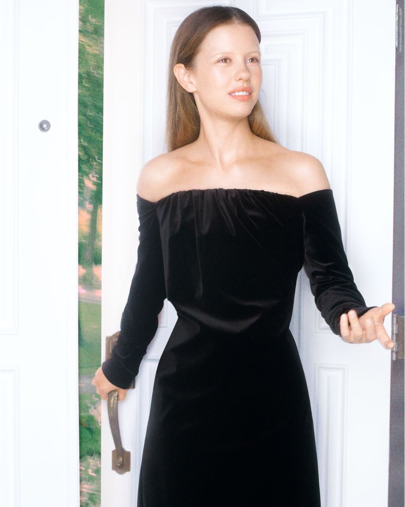 A woman in a black off-the-shoulder dress is opening a white door with a view of greenery outside, shot in natural lighting.
