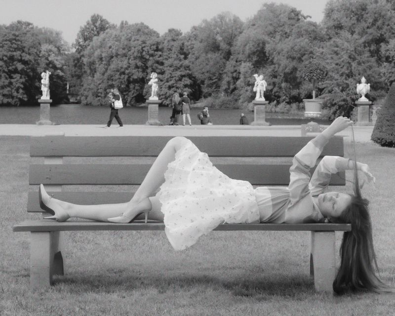 Monochromatic image of a woman reclining on a park bench wearing a dress and high heels, with trees and statues in the background.