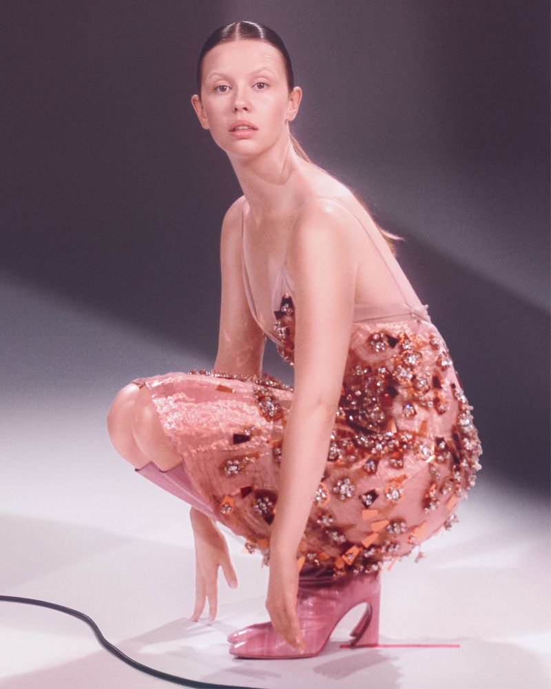 A slender adult woman in a crouched pose, wearing a pink embellished dress and matching heeled shoes, against a soft grey backdrop.