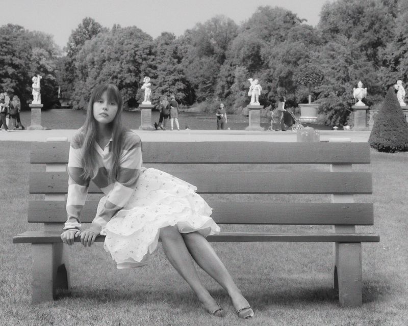 Black and white shot of a woman seated on a park bench wearing a striped shirt and floral dress in a grassy area with trees.