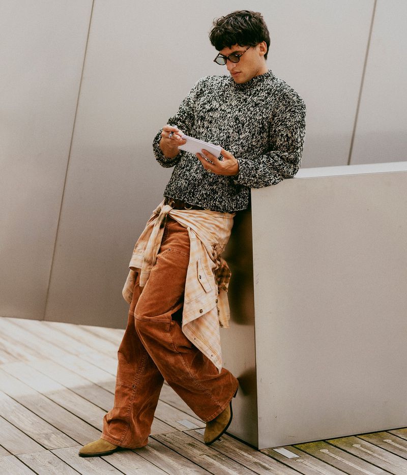 Man in a patterned sweater leans against a gray wall while holding a phone, wearing tan pants and tan suede boots in a casual style.