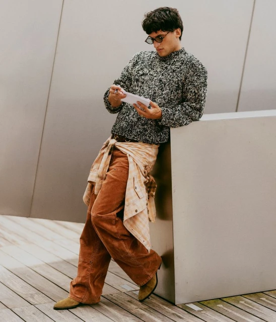 Man in a patterned sweater leans against a gray wall while holding a phone, wearing tan pants and tan suede boots in a casual style.