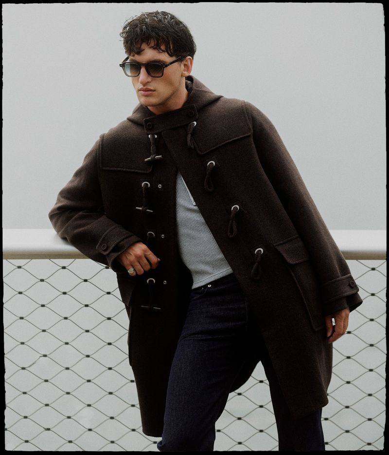 Man in a brown duffel coat and sunglasses poses outdoors by a metal fence, showcasing a blend of fashion and urban lifestyle.