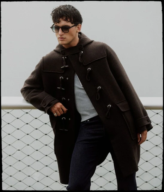 Man in a brown duffel coat and sunglasses poses outdoors by a metal fence, showcasing a blend of fashion and urban lifestyle.