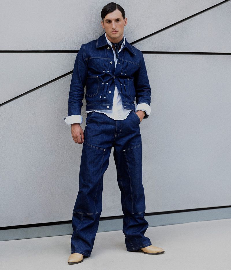 A male fashion model poses in a coordinating dark-wash denim jacket and wide-leg pants, with a white shirt, tan shoes, and patterned scarf.