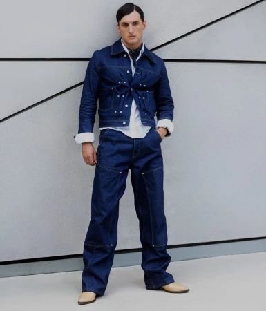 A male fashion model poses in a coordinating dark-wash denim jacket and wide-leg pants, with a white shirt, tan shoes, and patterned scarf.