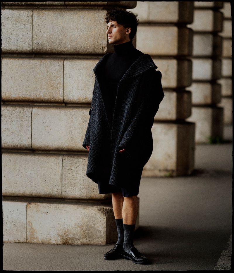 Man wearing a dark textured coat and black turtleneck standing against large stone columns in a fashion editorial.