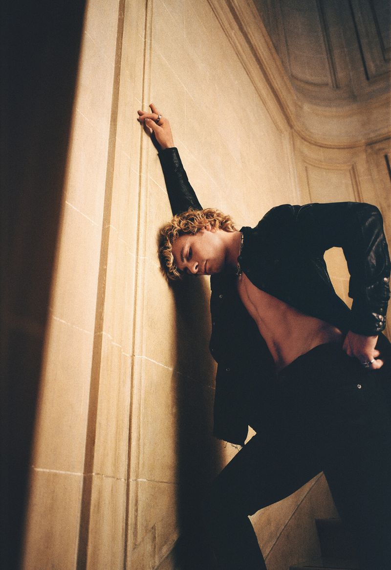 A slender adult man with curly blond hair poses against a wall indoors, wearing a dark jacket and pants, showcasing a fashionable aesthetic.
