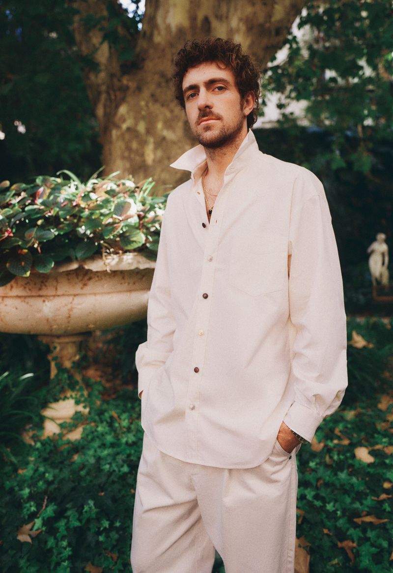 Portrait of a young man in a white linen shirt and trousers, standing in a lush outdoor garden setting with a stone planter.