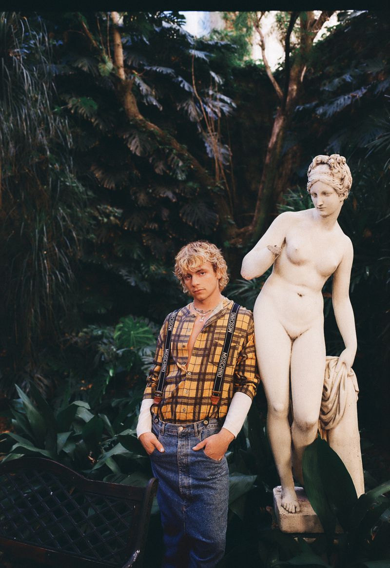 Man with blond hair and denim skirt poses next to nude statue in lush outdoor garden setting with a variety of green plants and trees.