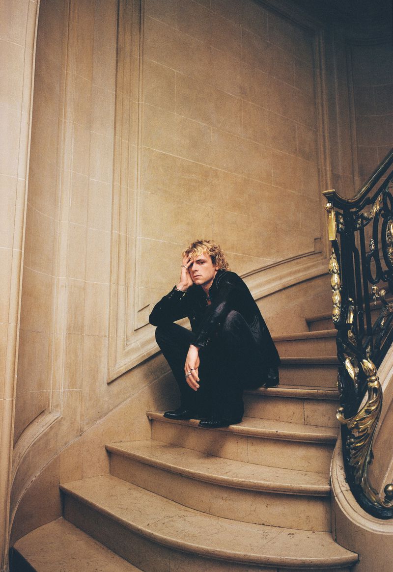 Man with blond hair sits on marble stairs indoors. He wears black clothes and poses with a hand near his face in a photo studio.