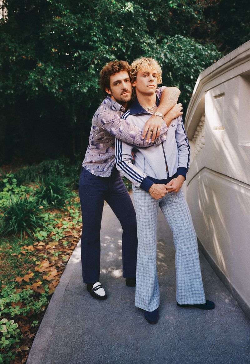Two men standing outdoors on a walkway by a building, one hugging the other, both wearing pants and shirts in a lifestyle shot.