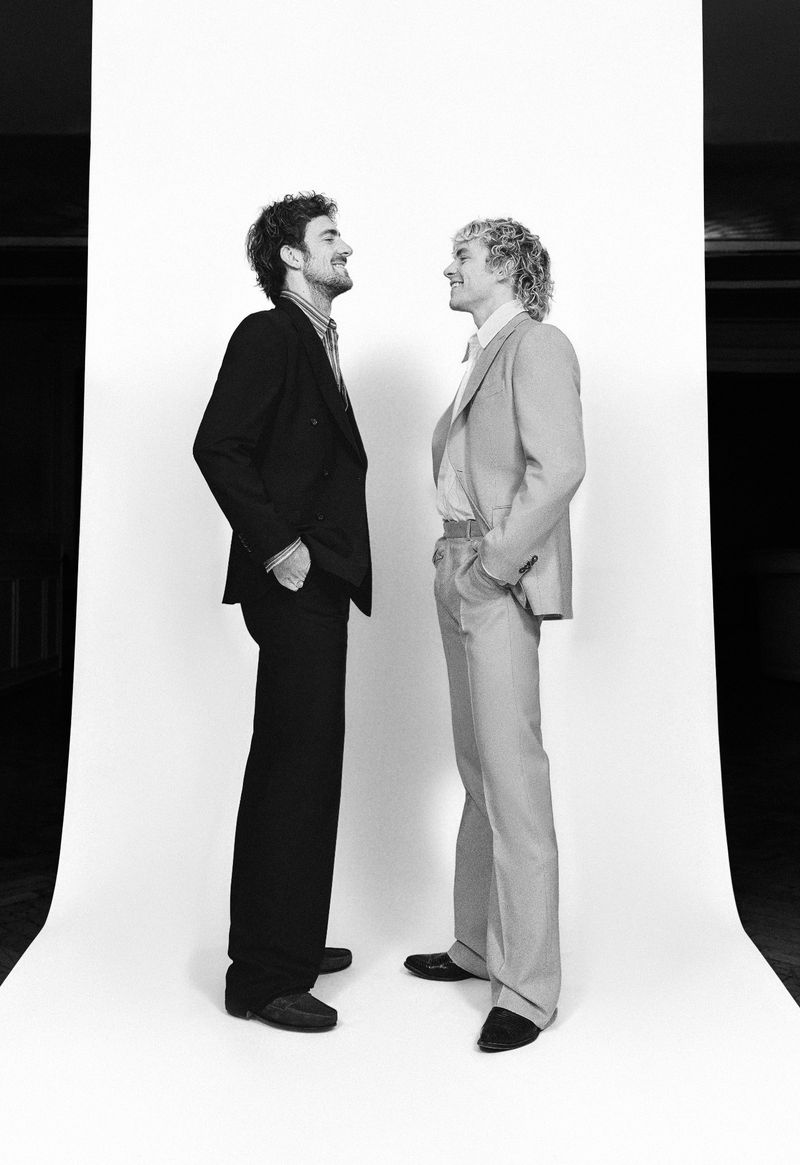 Two adult men in suits standing side by side in a photo studio, looking at each other, against a white backdrop in black and white.