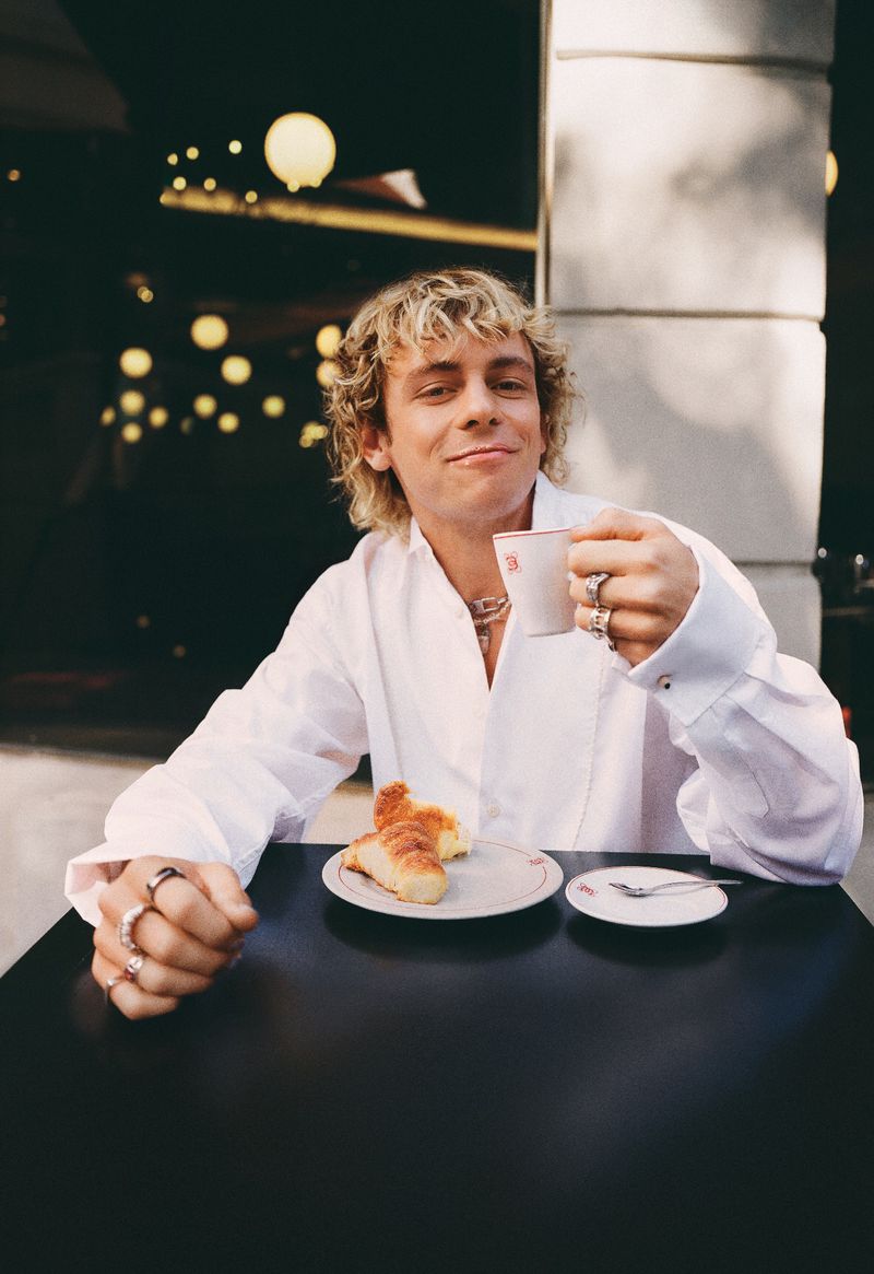 Young adult with blond hair sits at a table outside with a cup of coffee and croissants, wearing a white shirt and multiple rings.