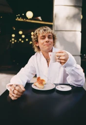 Ross Lynch seated at an outdoor café table wearing a white shirt and holding a small espresso cup.
